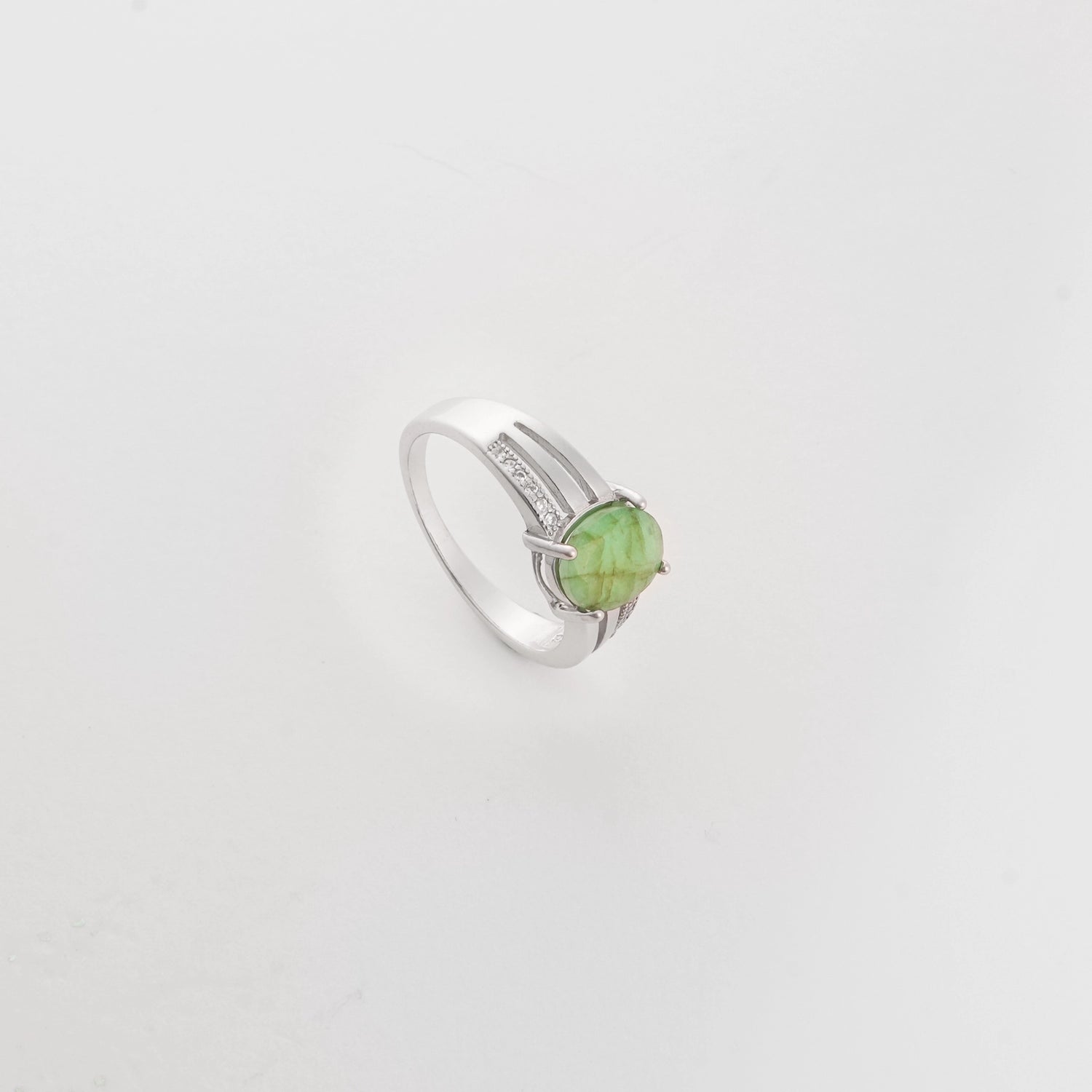 The Emerald Aura ring showcases a striking round green gemstone, secured in a sleek silver setting. Bright zircon stones line the split band, adding shimmer and balance.